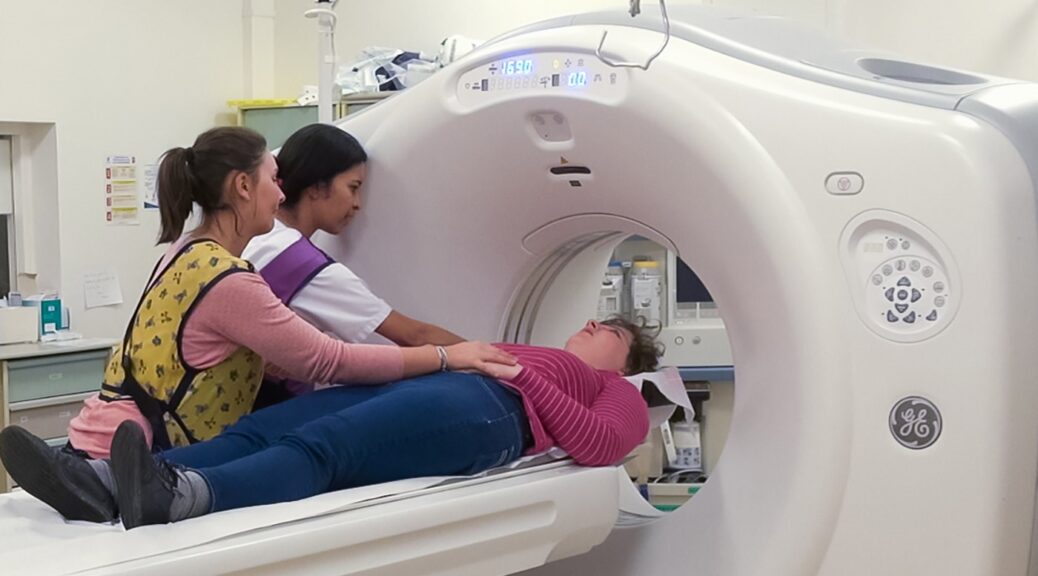 Une femme avec un trouble du spectre autistique est allongée pour effectuer un scanner lors d'une simulation de cet examen. Elle est accompagnée par une professionnelle de santé et par son aidante.
