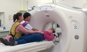 Une femme avec un trouble du spectre autistique est allongée pour effectuer un scanner lors d'une simulation de cet examen. Elle est accompagnée par une professionnelle de santé et par son aidante.